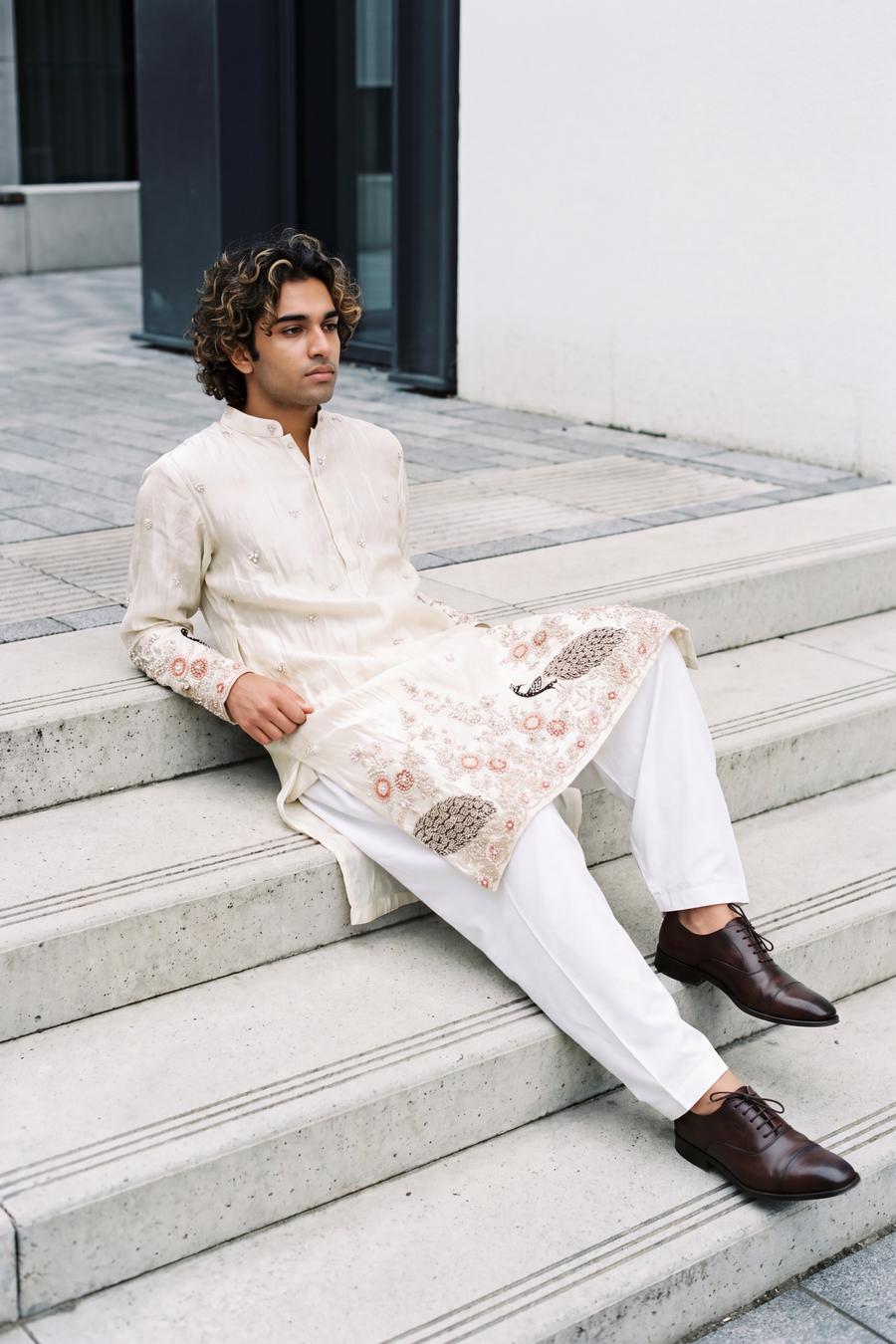 Ivory tissue silk kurta with peacock embroidered hem and white shalwar, model seated in a city setting