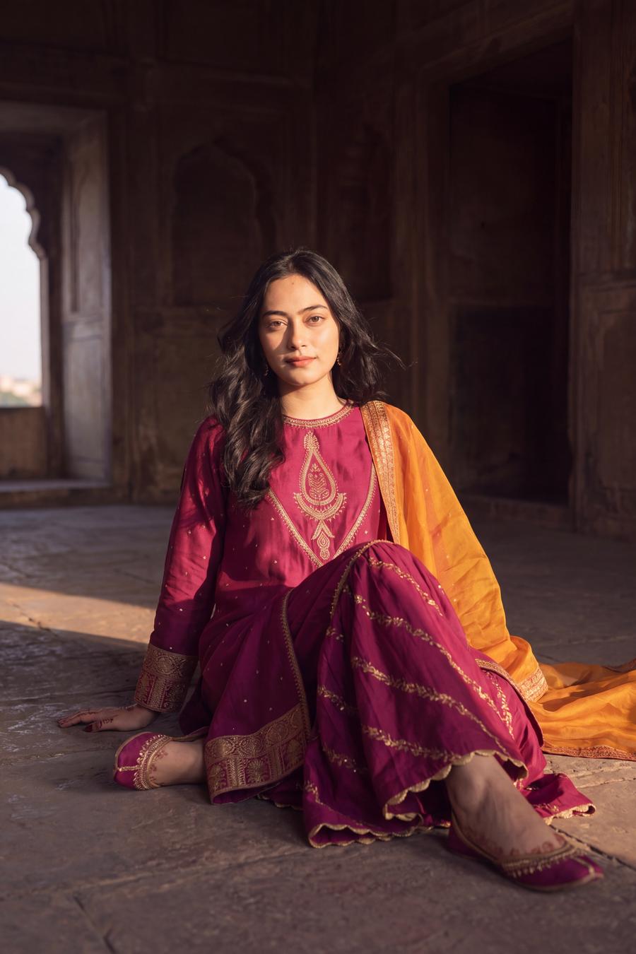 Magenta long shirt and sharara with marigold dupatta, seated on the floor with the sharara spread out