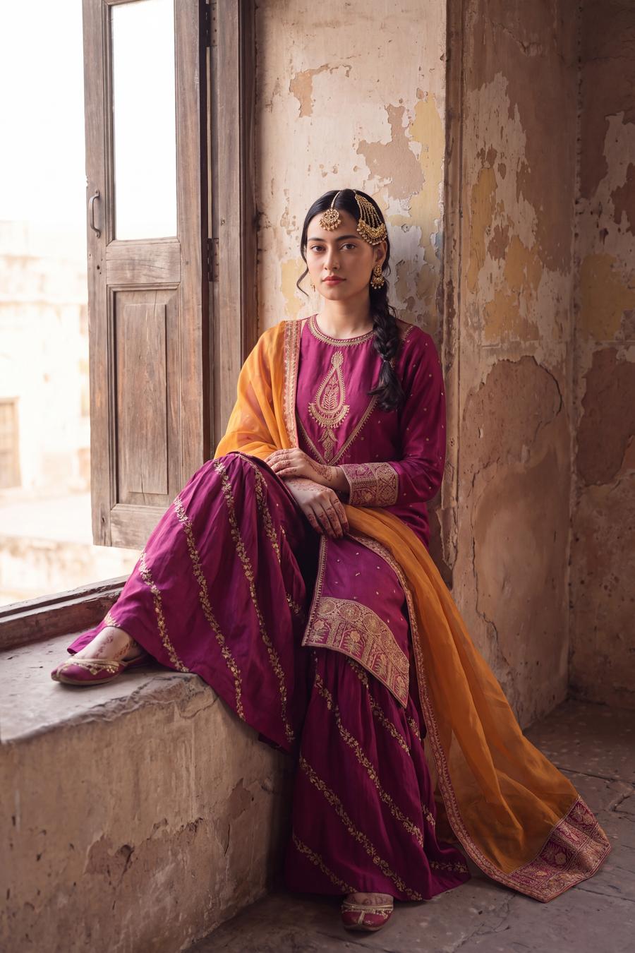 Magenta long shirt and sharara with marigold dupatta, seated by a window, styled with a side braid and pearl headpiece